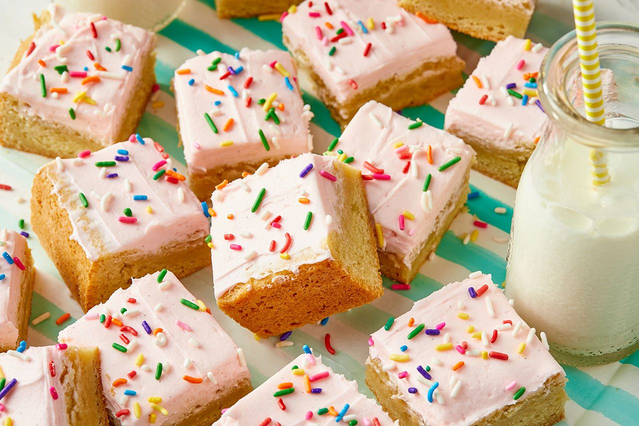 3/4 angle view shot of several square Lofthouse cookie bars topped with pale pink frosting and colorful rainbow sprinkles, arranged on a teal and white striped surface. A glass bottle of milk with a yellow and white striped straw is positioned on the right side of the frame;