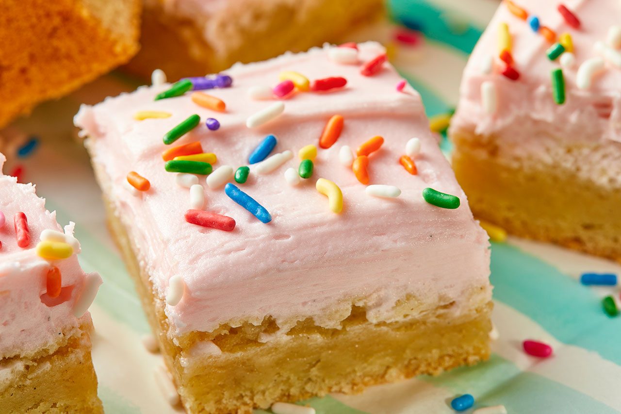 Close-up shot of a Lofthouse cookie bar topped with smooth pale pink frosting and colorful rainbow sprinkles, highlighting its soft, cakelike texture and thick layer of frosting, with additional frosted cookie bars softly blurred in the background;
