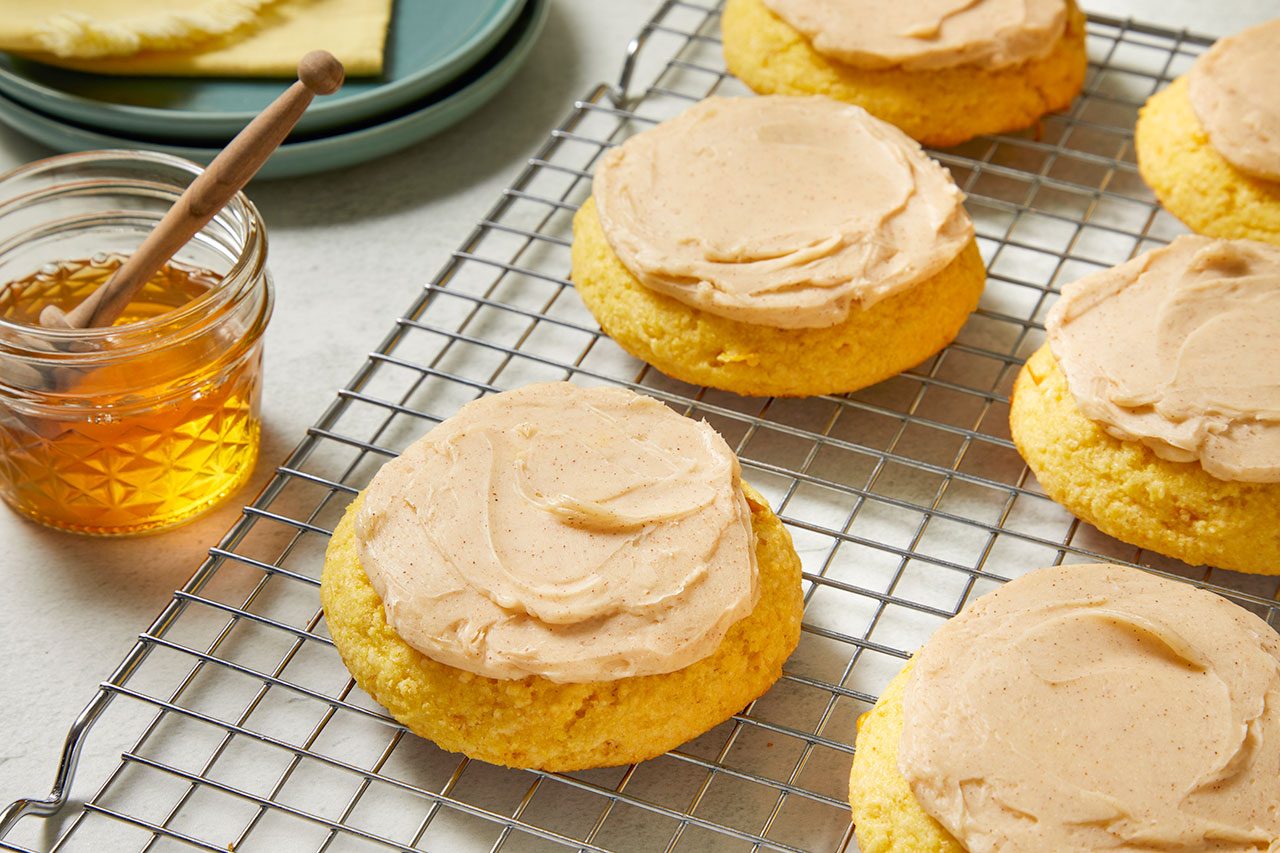3/4 angle view shot of frosted corn cookies arranged on a wire cooling rack, each topped with a smooth layer of creamy frosting, with a jar of honey and stacked plates in the background;