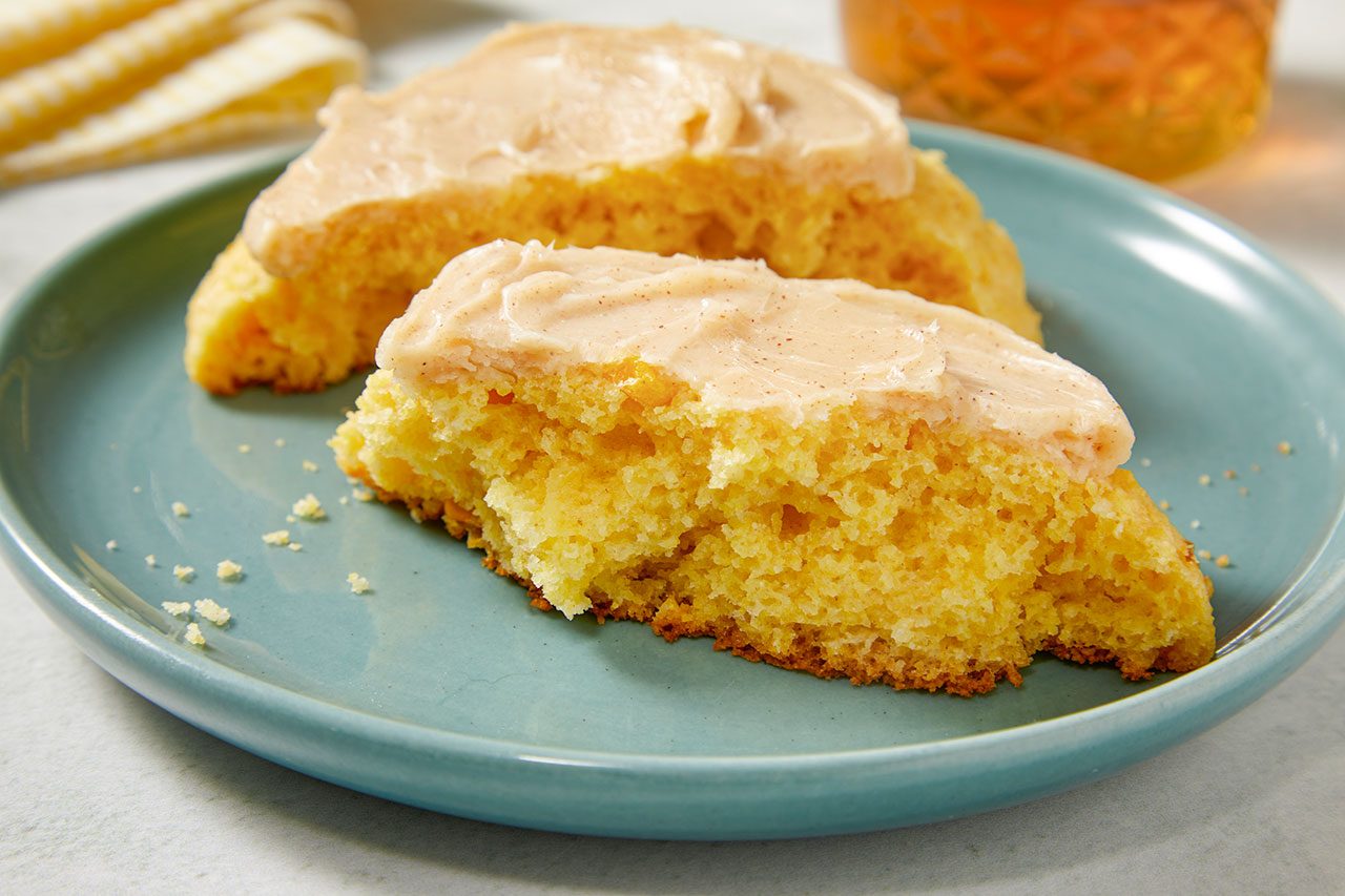 Close-up shot of a frosted corn cookie split in half on a blue plate, revealing its soft, tender crumb and topped with a smooth layer of creamy frosting, with a jar of honey softly blurred in the background;