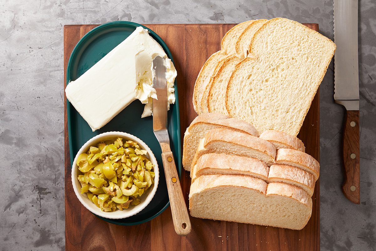 Sliced white bread on a wooden cutting board, a platter with a slab of cream cheese and a spreading knife, and a small bowl of chopped green olives with pimentos. A bread knife is beside the board.