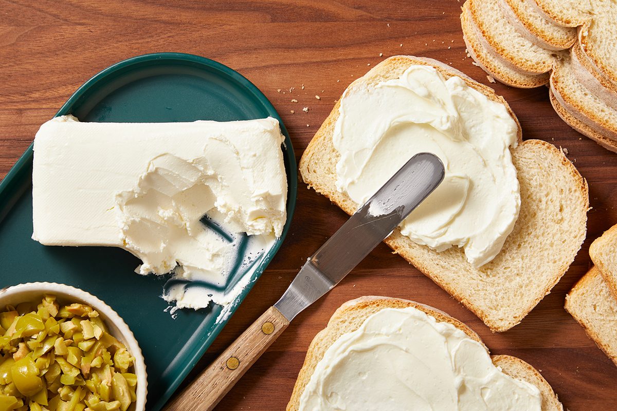 A knife spreads cream cheese on slices of bread, with a block of cream cheese on a green plate and a small bowl of chopped pickles nearby, all on a wooden surface.