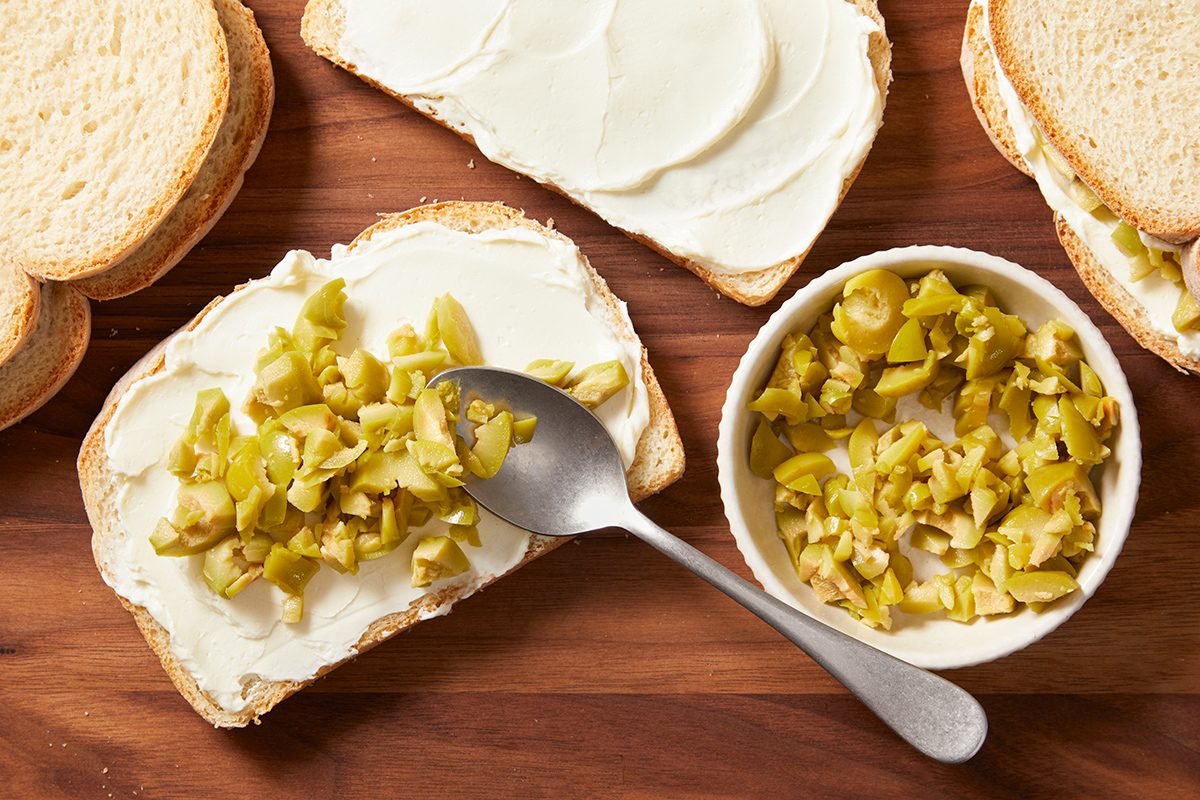 Slices of bread on a wooden surface, one topped with cream cheese and chopped green olives. A bowl of chopped olives with a spoon sits nearby.