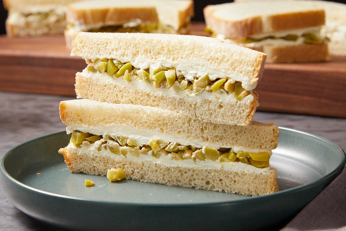 Two sandwich halves with white bread, a creamy filling, and chopped green olives are stacked on a gray plate. More similar sandwiches are visible in the background on a wooden board.