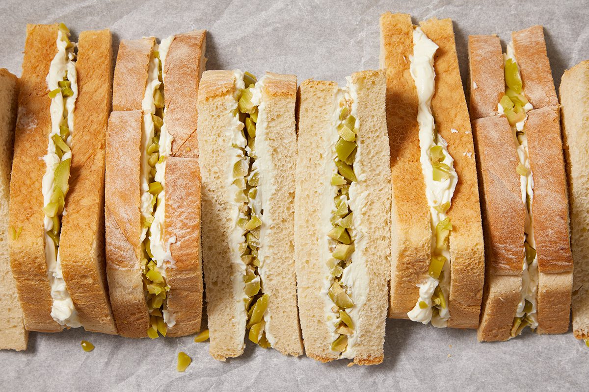 Several neatly arranged sandwiches made with white bread, filled with a mixture of chopped green pickles and a creamy spread, are placed side by side on parchment paper.