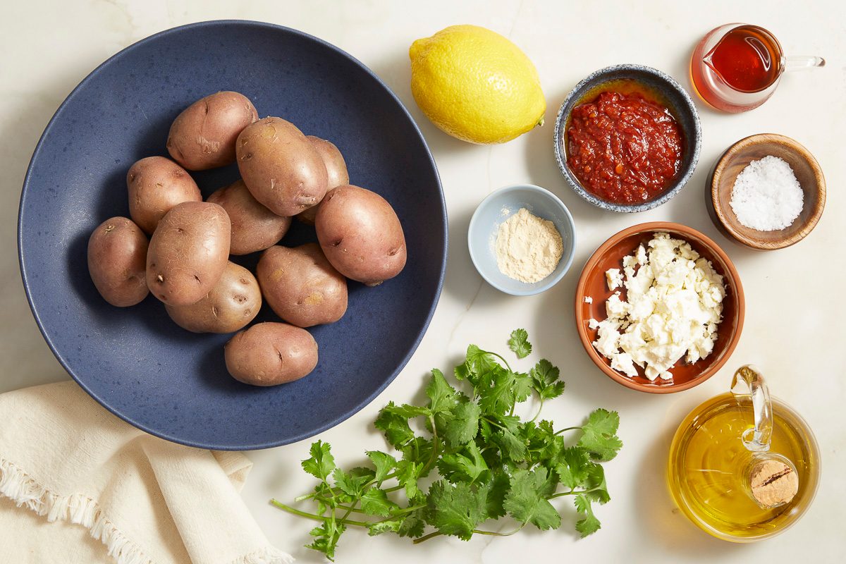 Overhead shot of potatoes alongside bowls of olive oil, harissa paste, hot honey, crumbled feta, garlic powder, salt, lemon, fresh cilantro, and seasonings.