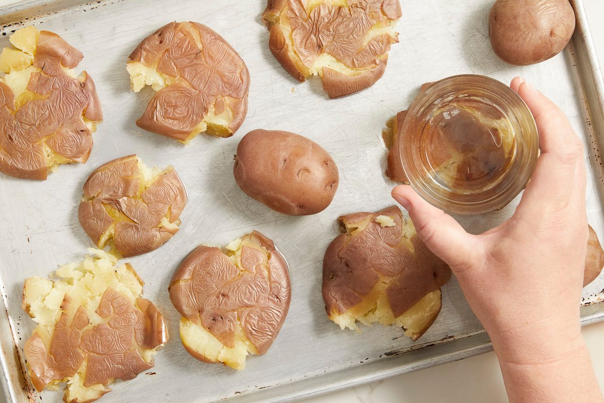 Overhead shot of smashed baby potatoes arranged on a parchment-lined baking sheet as a hand brushes them with a glossy harissa honey mixture using a small glass bowl;