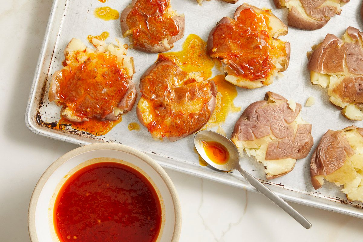 Overhead shot of smashed baby potatoes on a parchment-lined baking sheet, generously brushed with a glossy harissa honey glaze; A bowl of extra glaze and a spoon rest nearby, while additional potatoes await coating.