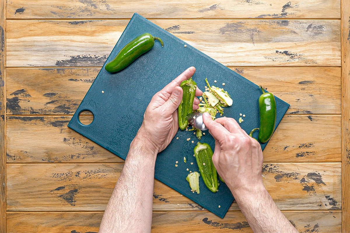 Overhead shot of a person scoops seeds out of a jalapeño pepper with a spoon on a cutting board more jalapeños are nearby on the wooden table