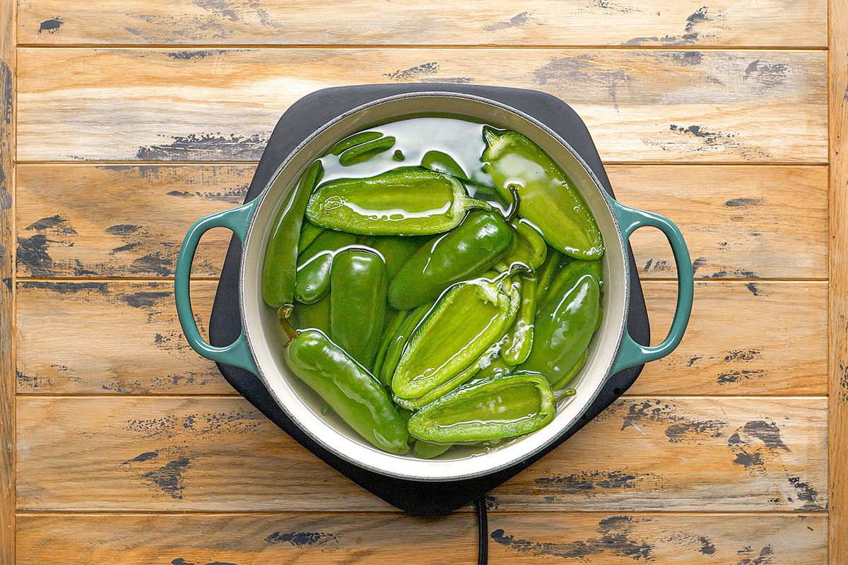 Overhead shot of a pot of whole green jalapeño peppers is boiling in water on a wooden surface The steam rises as the peppers cook together