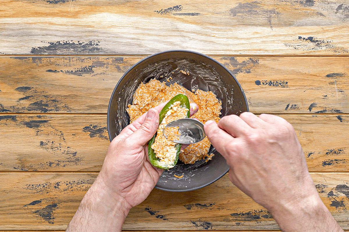 Overhead shot of a person spoons a creamy orange mixture into half a jalapeño pepper on a rustic wooden surface with more filling in a nearby bowl