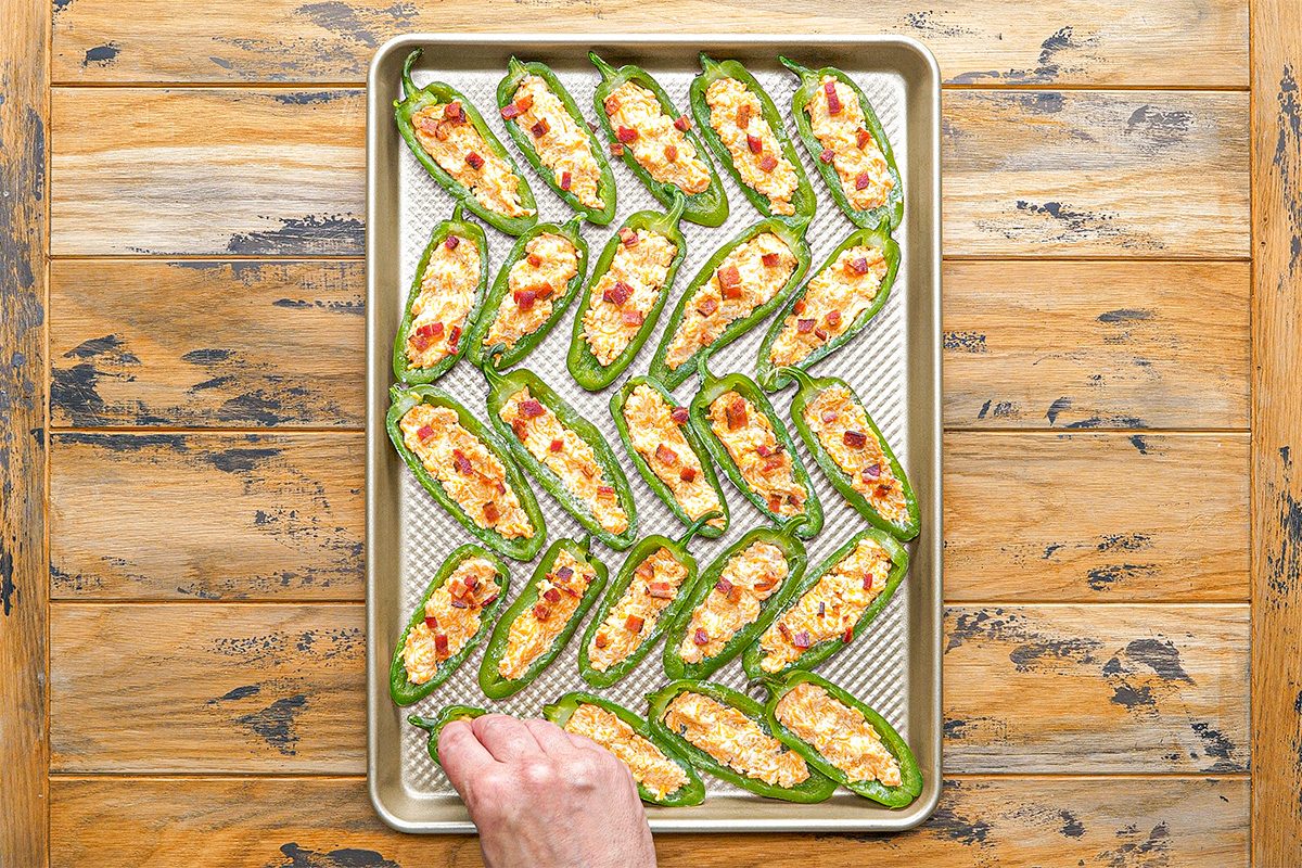 Overhead shot of a hand reaches for a jalapeño half stuffed with creamy filling and bacon pieces on a baking sheet with more stuffed jalapeños on wood