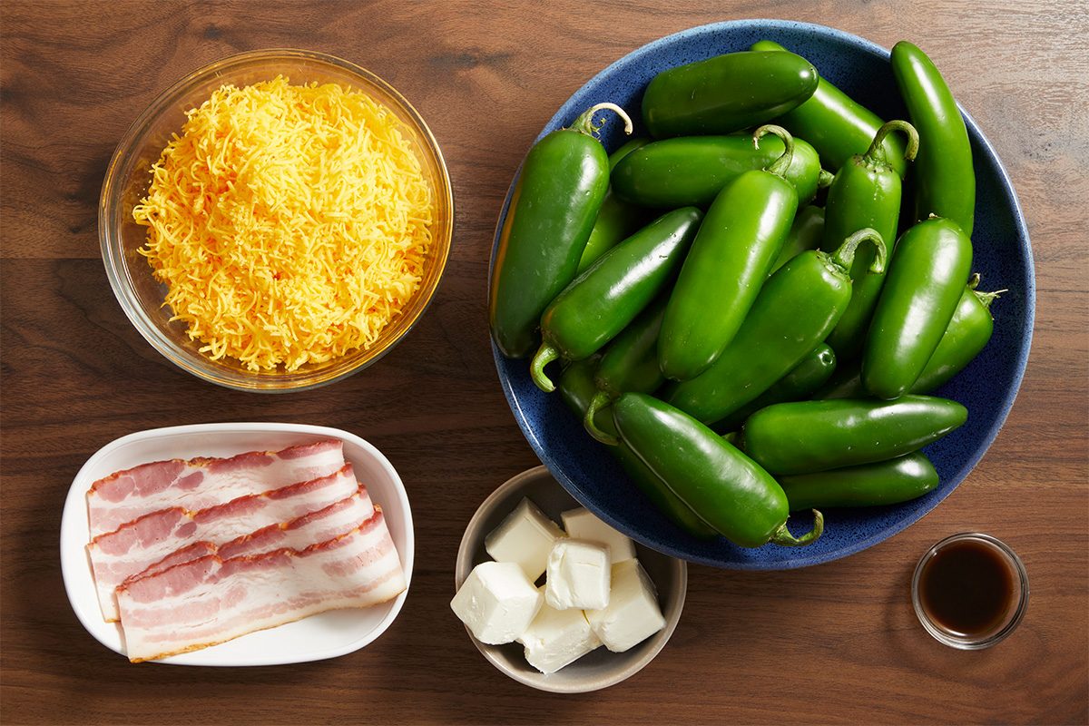 Overhead shot of raw ingredients including shredded cheddar cheese, whole jalapeños in a bowl, strips of raw bacon, cubed cream cheese, and seasoning on a wooden surface.