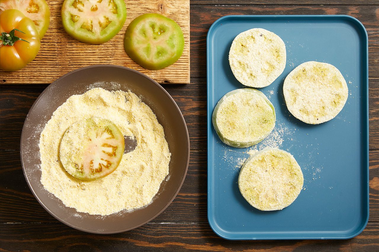 Overhead horizontal image of a step in preparing Southern Fried BLTs, showing a green tomato slice being coated in a cornmeal mixture within a brown bowl. Several other coated green tomato slices are neatly arranged on a blue rectangular tray, while additional sliced and one whole green tomato rest on a wooden cutting board.