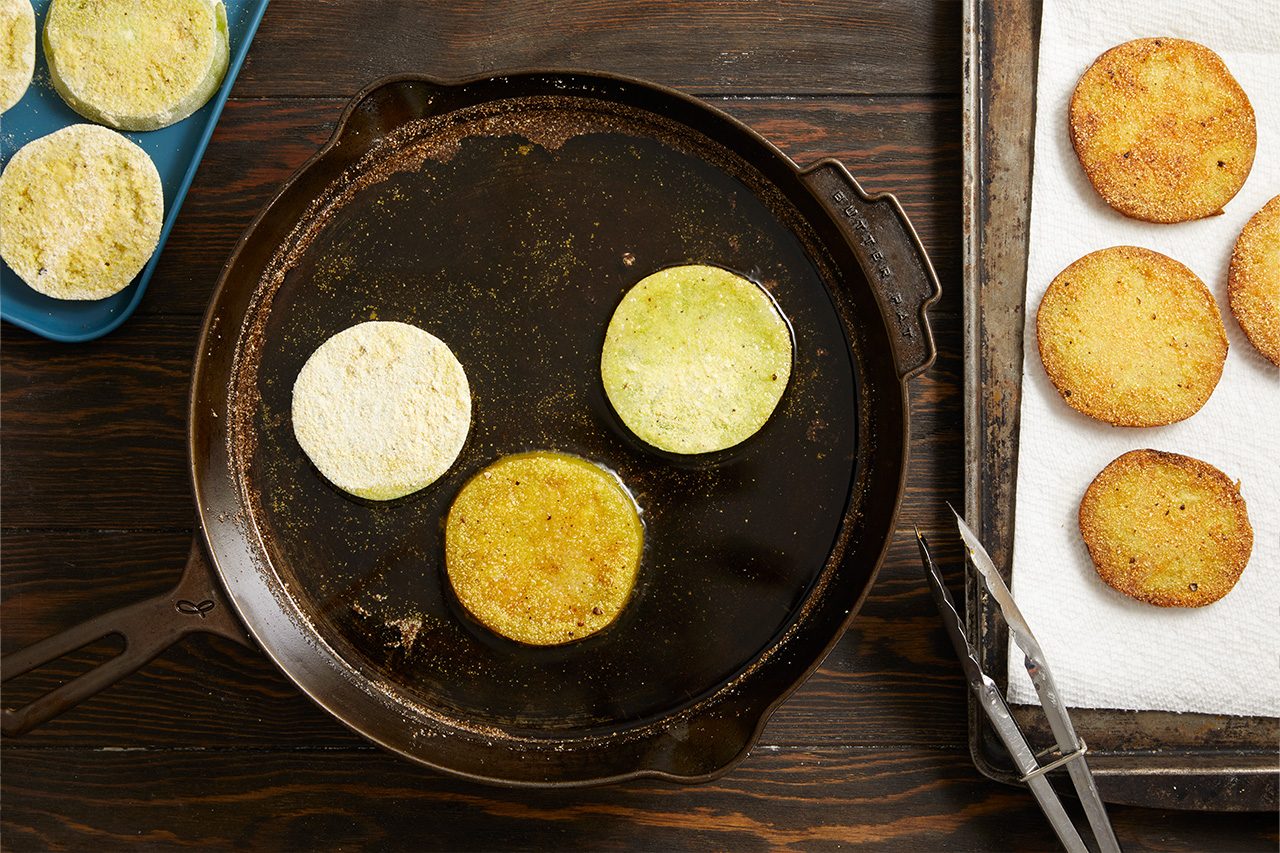 Overhead horizontal image of green tomato slices being fried in a cast iron skillet. Unfried, coated slices wait on a blue platter, while others sizzle in hot oil, and some golden brown, fried slices drain on paper towels.