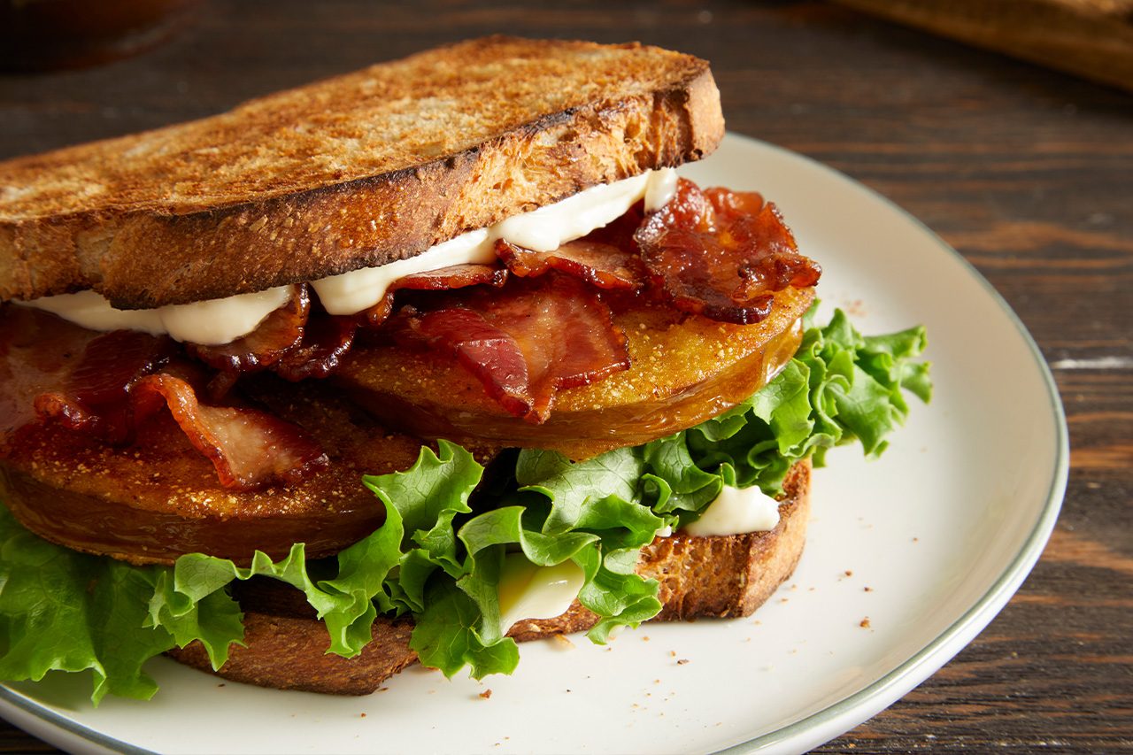 Eye Level horizontal image of a Southern Fried BLT sandwich on a white plate. The sandwich features toasted whole wheat bread, crispy cooked bacon strips, fried green tomato slices, fresh iceberg lettuce leaves, and creamy mayonnaise.