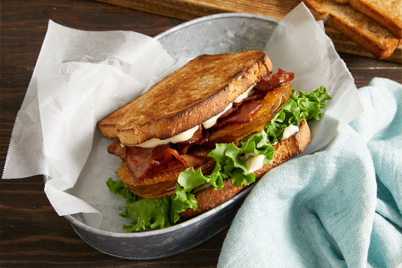 Eye Level horizontal image of a Southern Fried BLT sandwich nestled in a metal tin lined with white parchment paper. The sandwich is made with toasted whole wheat bread, fried green tomatoes, crispy bacon, iceberg lettuce, and a generous spread of mayonnaise, with a light blue napkin visible beside it.