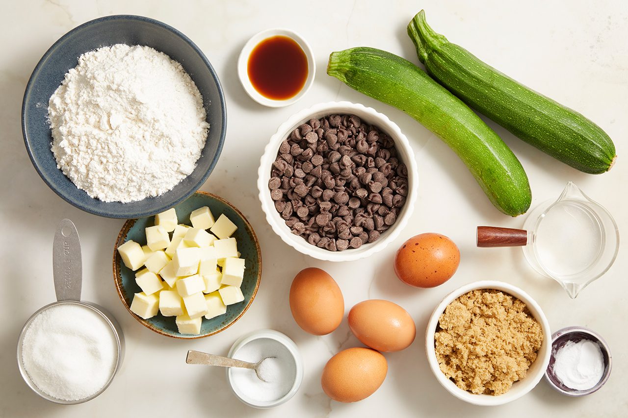 Baking ingredients including flour, sugar, baking soda, brown sugar, butter, eggs, chocolate chips, vanilla, milk, and two whole zucchinis are arranged on a countertop.