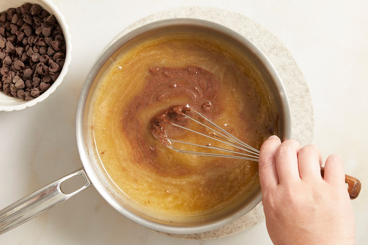 A hand uses a whisk to stir melted chocolate into a saucepan of brown batter, with a bowl of chocolate chips nearby on a white surface.