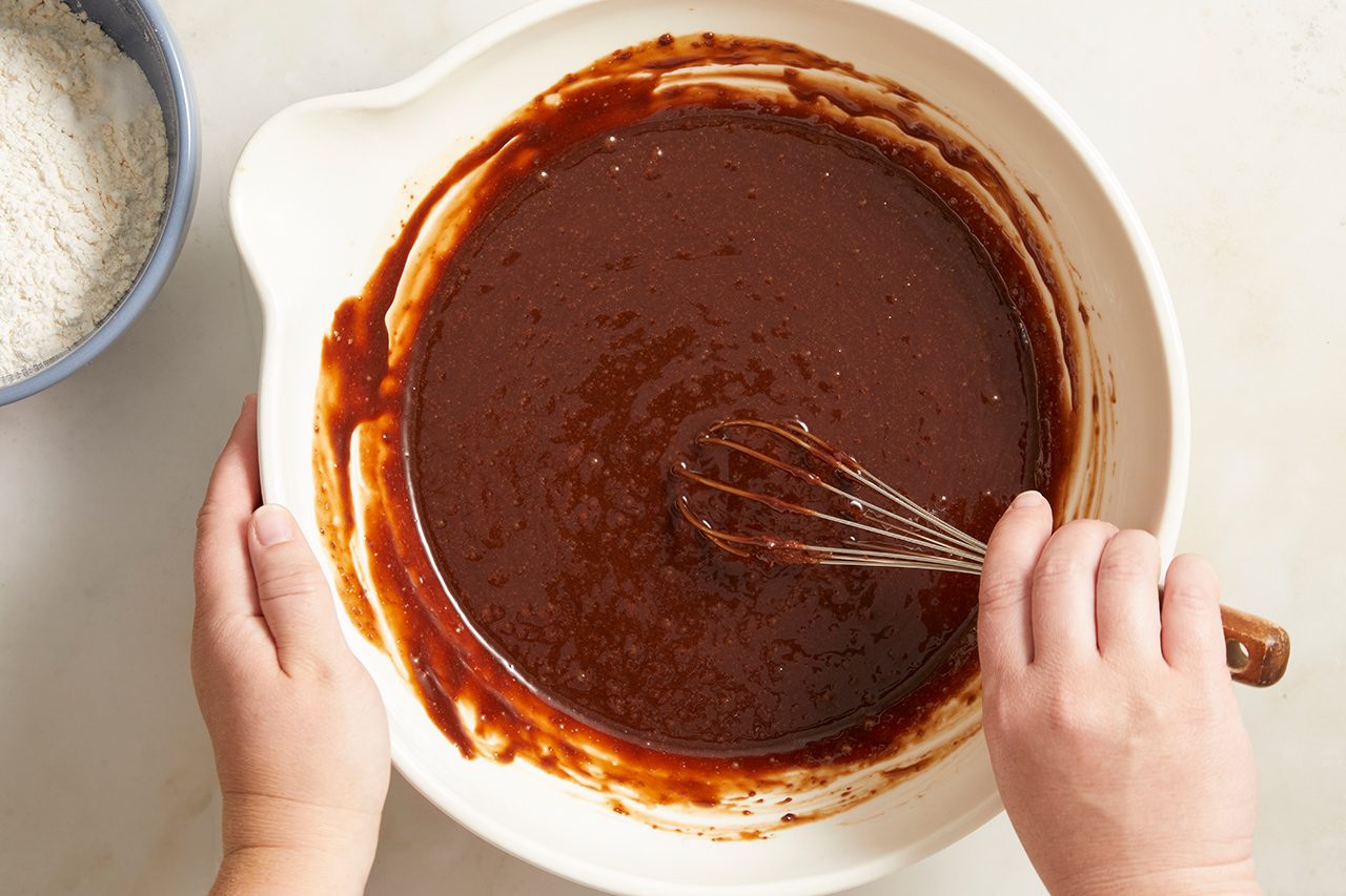 A person uses a whisk to mix chocolate batter in a white bowl, with a smaller bowl of flour visible nearby on a light surface.