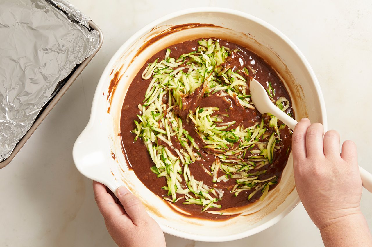 Hands mix shredded zucchini into chocolate batter in a white bowl with a spatula. A foil-lined baking pan sits nearby on a light countertop.