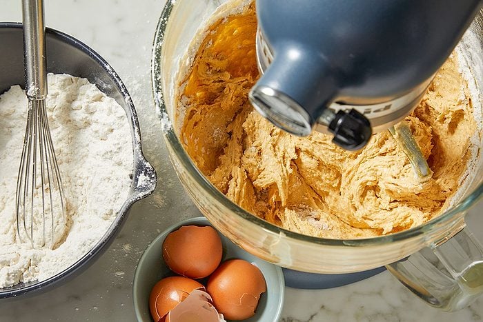 Overhead horizontal image of cookie dough being mixed in a dark blue stand mixer. A black bowl of flour with a whisk rests beside the mixer, and a small blue bowl holds brown eggshells and whole eggs.