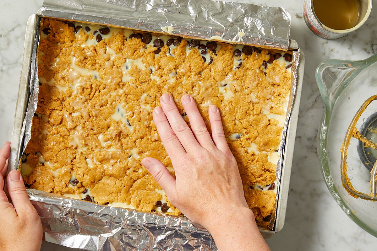 Overhead horizontal image of a hand pressing down a crumbly dough topping into a foil-lined baking pan. The pan contains chocolate chips and a milky liquid, likely sweetened condensed milk, beneath the topping. A mixing bowl with a paddle attachment and a can are visible in the background.