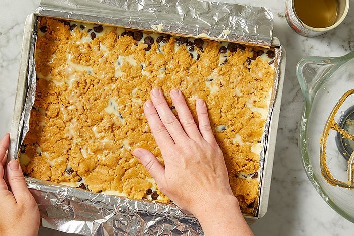Overhead horizontal image of a hand pressing down a crumbly dough topping into a foil-lined baking pan. The pan contains chocolate chips and a milky liquid, likely sweetened condensed milk, beneath the topping. A mixing bowl with a paddle attachment and a can are visible in the background.