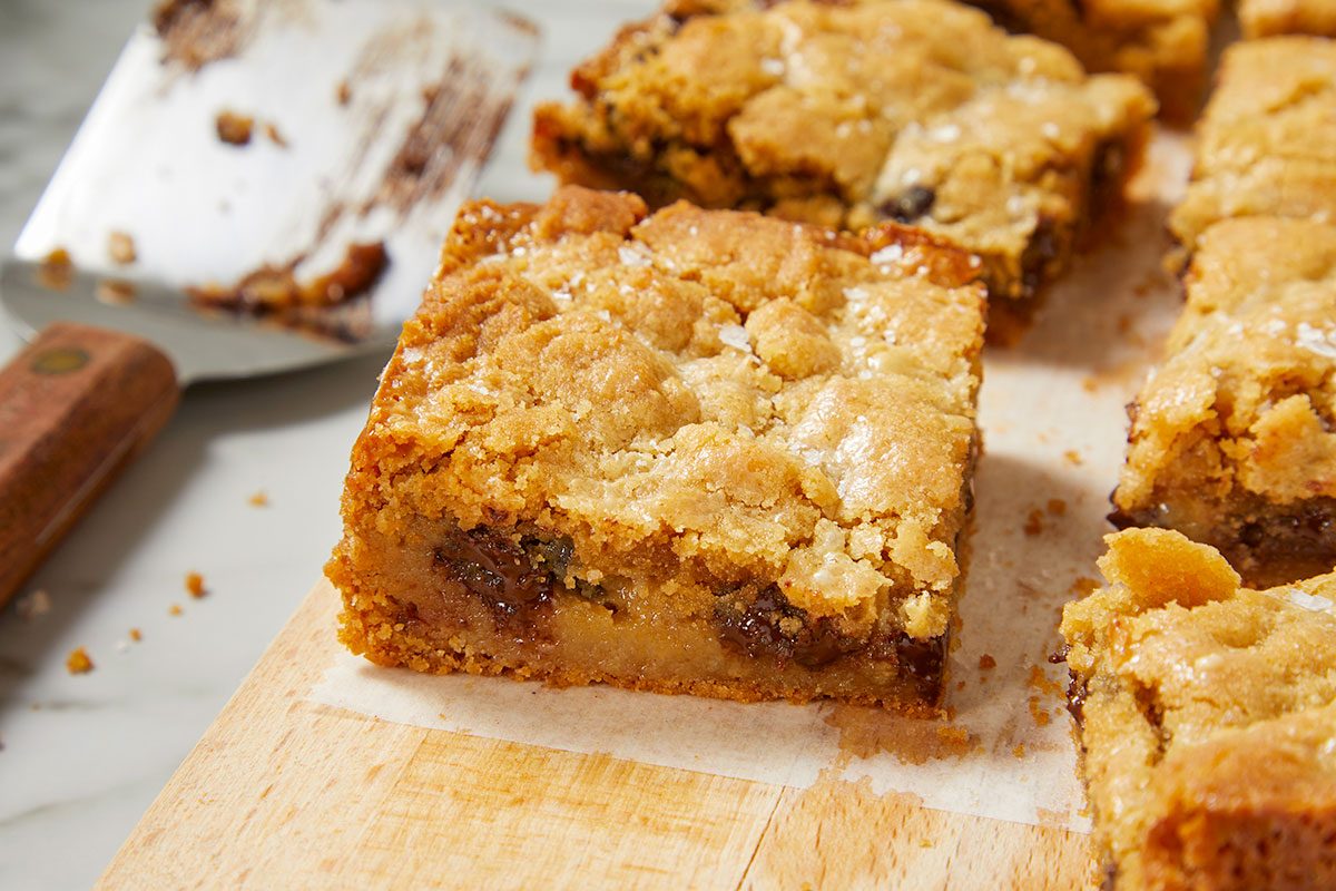 Eye Level horizontal image of several freshly baked blondies or dessert bars arranged on a wooden cutting board lined with parchment paper. Each bar features a golden-brown, crumbly top, sprinkled with coarse salt, revealing a gooey interior with visible chocolate chips. A used metal spatula is blurred in the background.