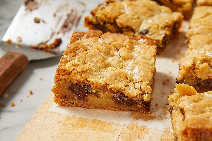 Eye Level horizontal image of several freshly baked blondies or dessert bars arranged on a wooden cutting board lined with parchment paper. Each bar features a golden-brown, crumbly top, sprinkled with coarse salt, revealing a gooey interior with visible chocolate chips. A used metal spatula is blurred in the background.
