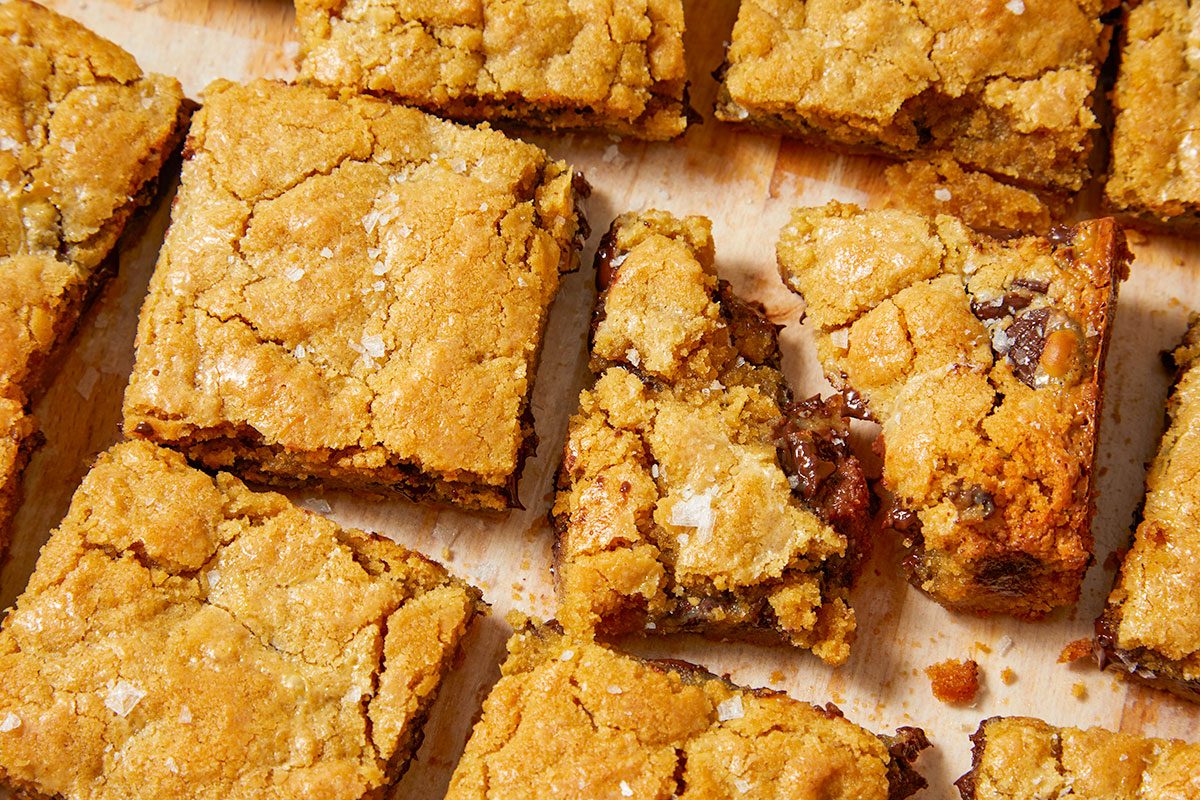Overhead horizontal image of golden-brown cookie bars, some broken, scattered on a light surface. The bars are speckled with flaky sea salt and reveal melty chocolate chips within their soft, chewy texture.