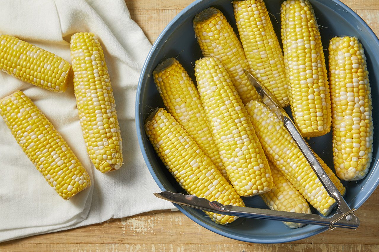 A blue bowl filled with several ears of raw corn on the cob, with metal tongs resting on top. More corn cobs sit on a white cloth nearby, all on a wooden surface.