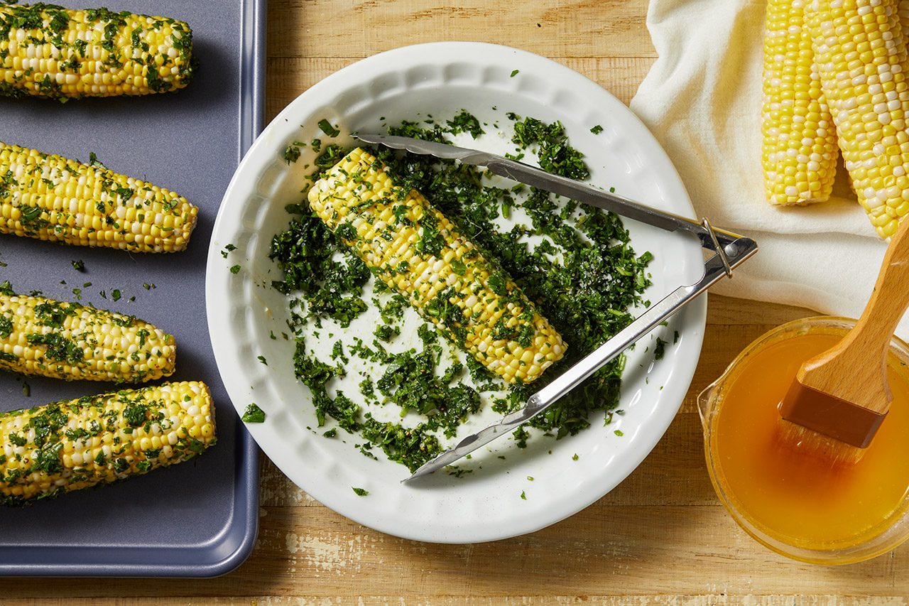Corn on the cob is being coated in chopped herbs with tongs in a white dish. Nearby are more ears of corn, a baking tray, a cloth, and a bowl of melted butter with a pastry brush.