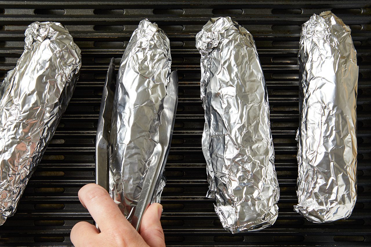 Four ears of corn wrapped in aluminum foil are on a grill grate. A hand holding tongs is turning one of the foil-wrapped corncobs.