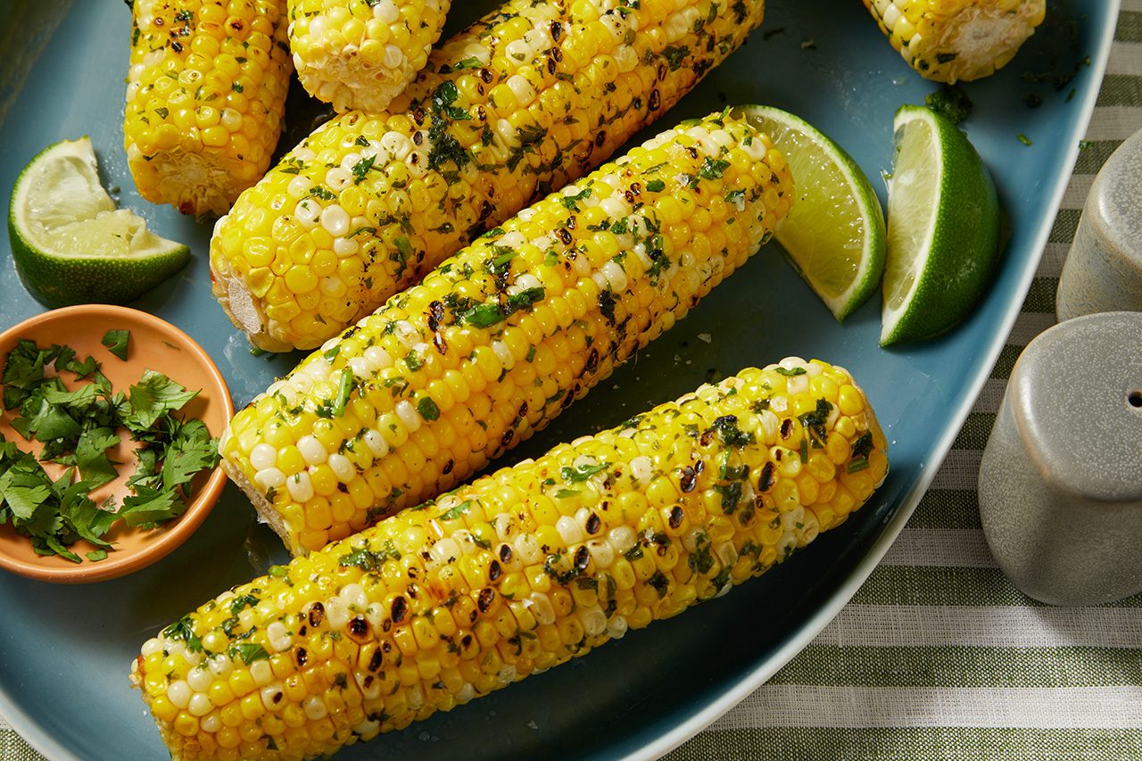 Grilled corn on the cob garnished with chopped herbs on a blue plate, served with lime wedges and a small dish of extra herbs.