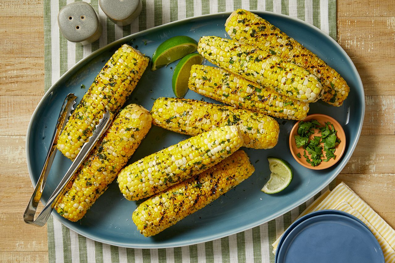 Grilled corn on the cob with herbs is arranged on a blue platter. Lime wedges, a small bowl of chopped herbs, salt and pepper shakers, and metal tongs are also on the table.