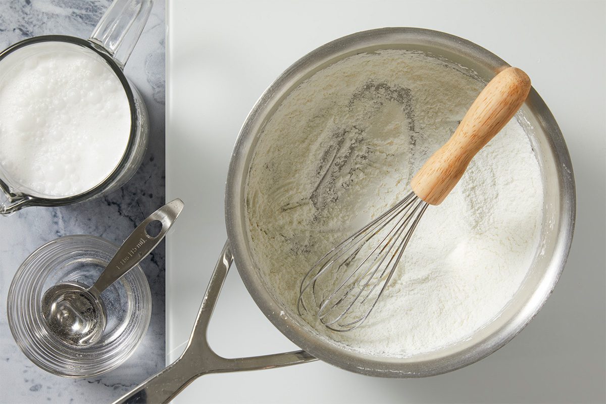 Overhead shot of dry ingredients being whisked together in a saucepan, with a measuring cup of milk and a small bowl of oil on a light countertop;