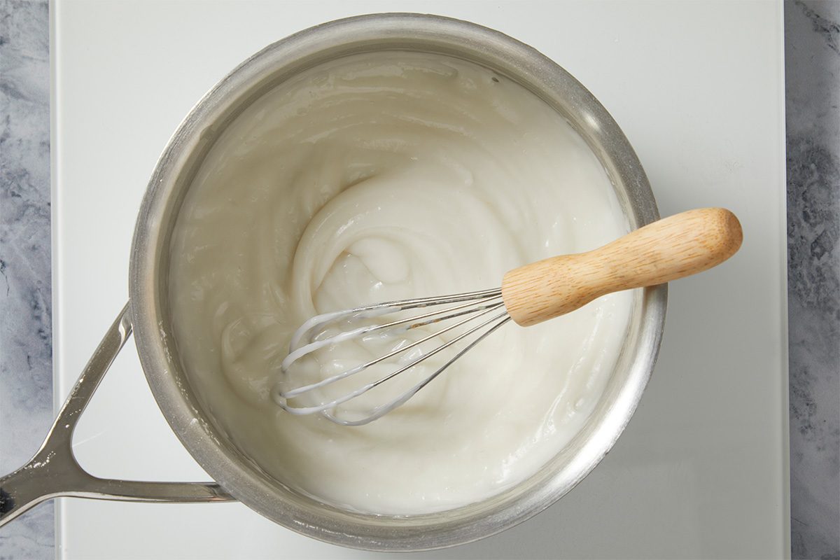 Overhead shot of a saucepan filled with smooth white sauce being whisked on a light countertop, highlighting its creamy, glossy texture;