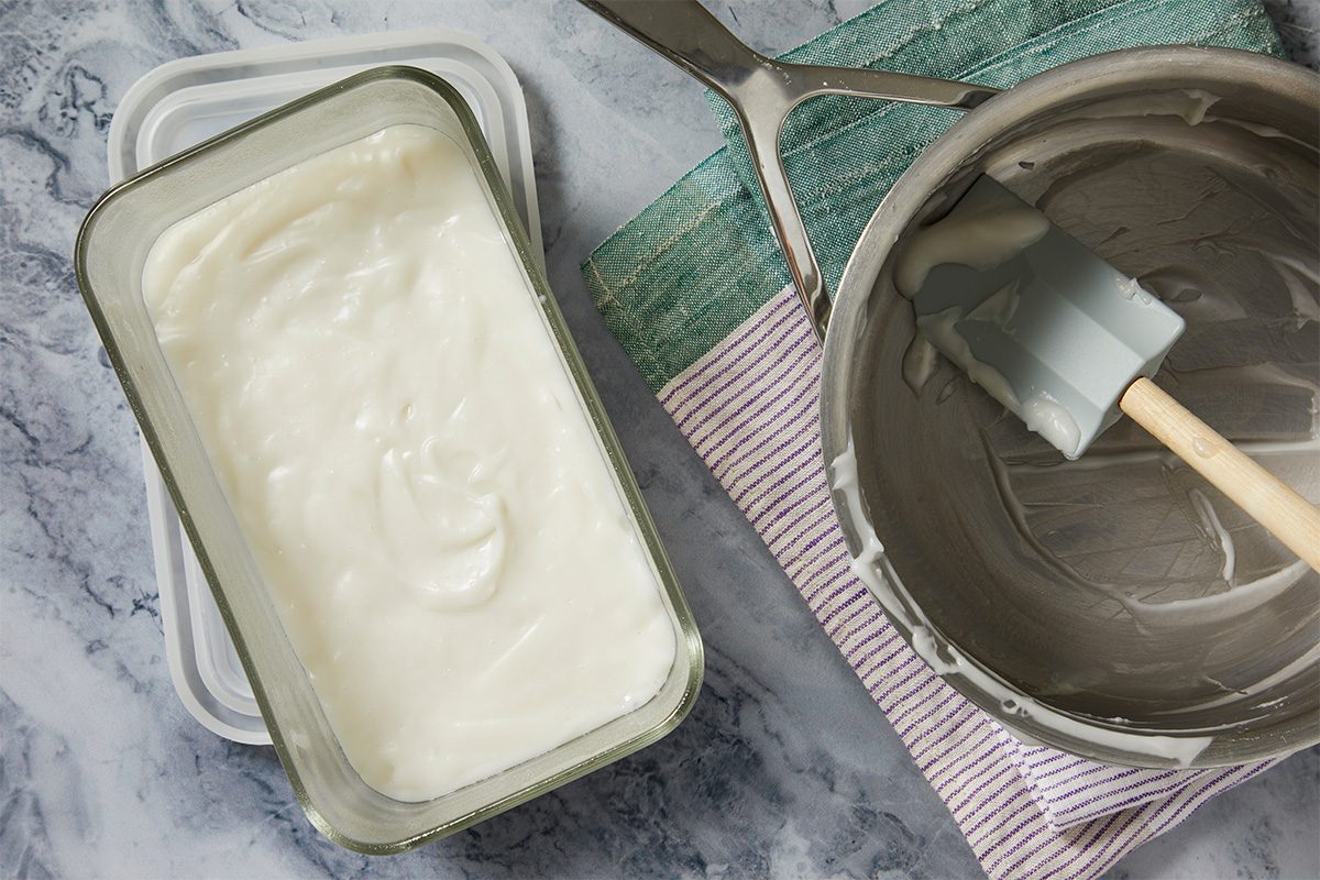 Overhead shot of freshly prepared creamy mixture spread in a loaf pan beside an empty saucepan with a spatula, set on a marble countertop with a kitchen towel;