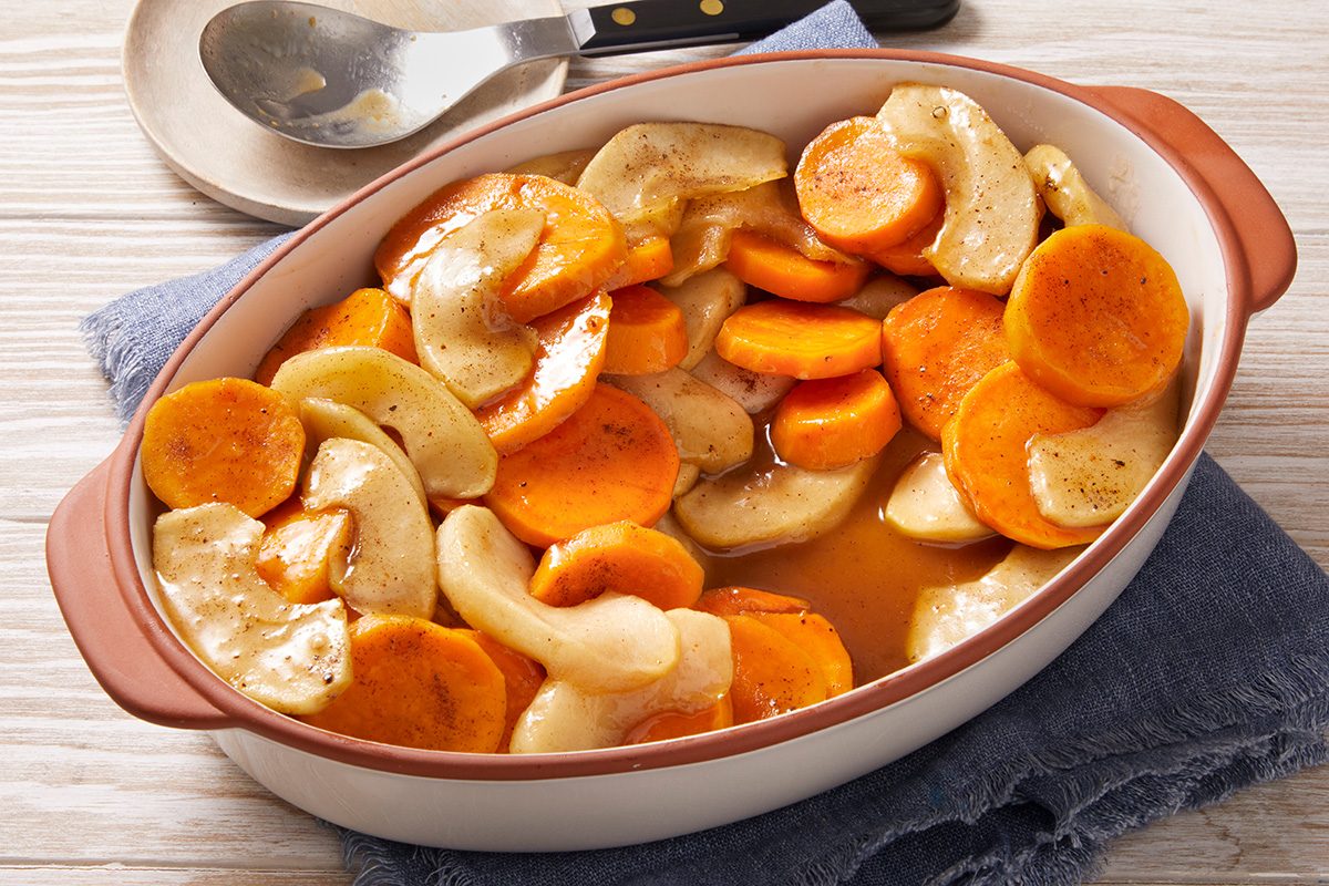 Oval baking dish filled with sliced sweet potatoes and apples in a light sauce, placed on a blue cloth with a spoon and plate in the background.