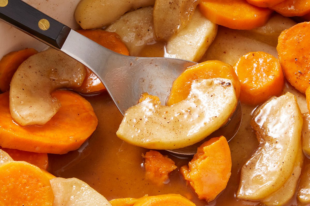 A close-up of a spoon scooping glazed sweet potato and apple slices in a spiced syrup, showing the tender, cooked pieces and glossy sauce.