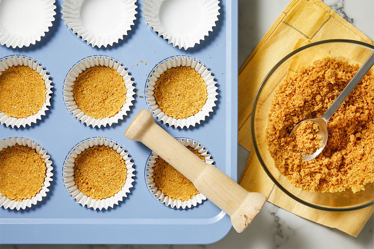 Overhead horizontal image of a blue muffin tin with white cupcake liners, some filled with a crumb crust, and a wooden tamper resting in one. A glass bowl of crumb mixture with a spoon sits on a yellow napkin beside the tin.