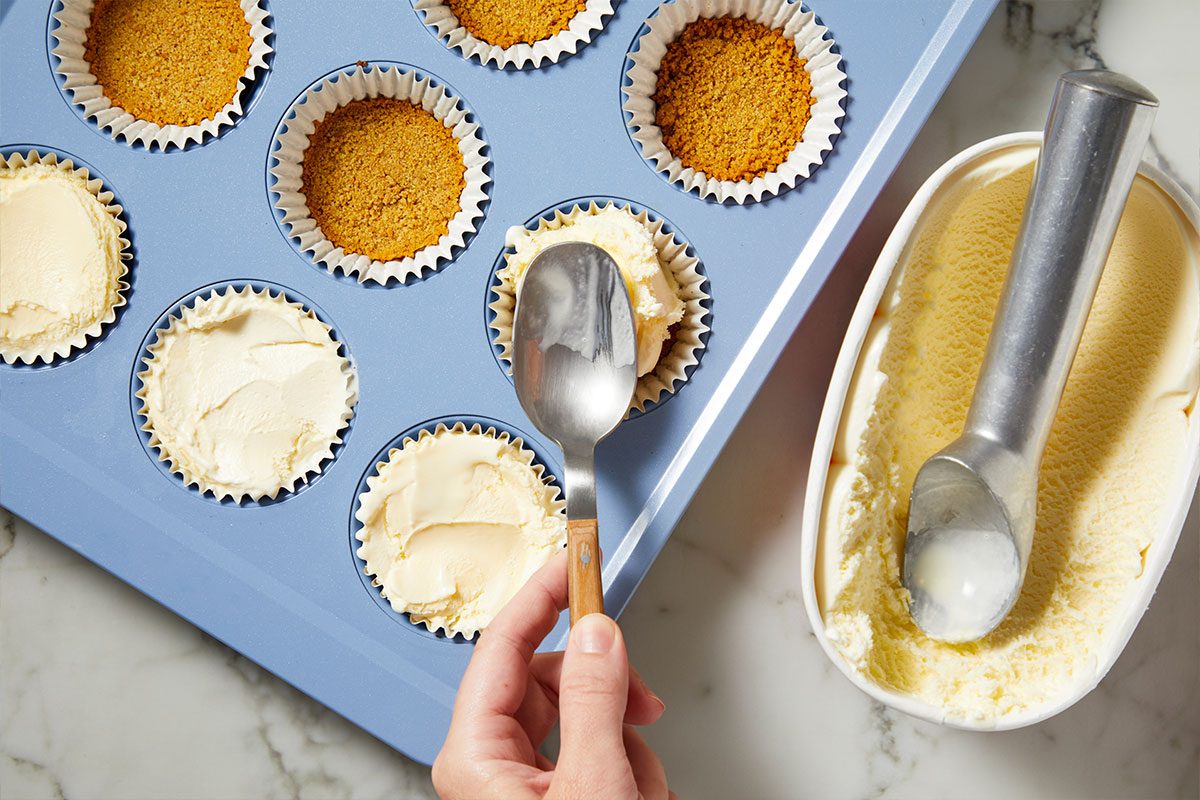 Overhead horizontal image of a hand using a spoon to press vanilla ice cream into a cupcake liner within a blue muffin tin. Several other liners are visible, some containing a golden-brown graham cracker crust and others already filled with ice cream. A container of vanilla ice cream with an ice cream scoop sits nearby on a marble surface.