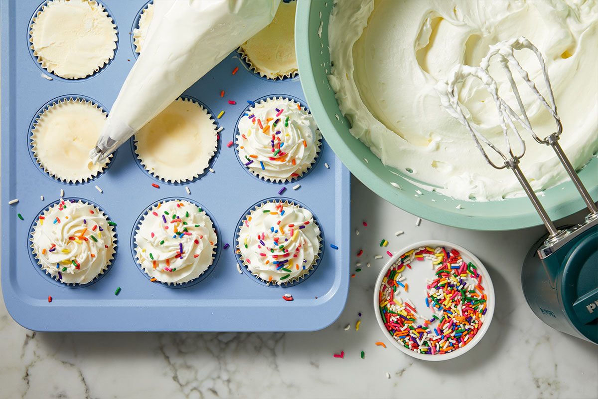 Overhead horizontal image of cupcakes being frosted and decorated. A piping bag is used to frost a cupcake in a blue muffin pan, while other frosted cupcakes are topped with colorful sprinkles. A light green bowl holds whipped cream with an electric mixer submerged, and a small dish of sprinkles sits nearby on a marble surface.