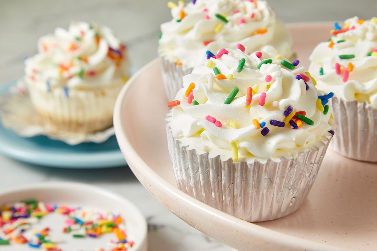 Eye-level horizontal image of several cupcakes on a pink cake stand, topped with swirls of white frosting and colorful sprinkles. A blurred cupcake is visible on a blue plate in the background, alongside a small white dish filled with loose sprinkles in the foreground.