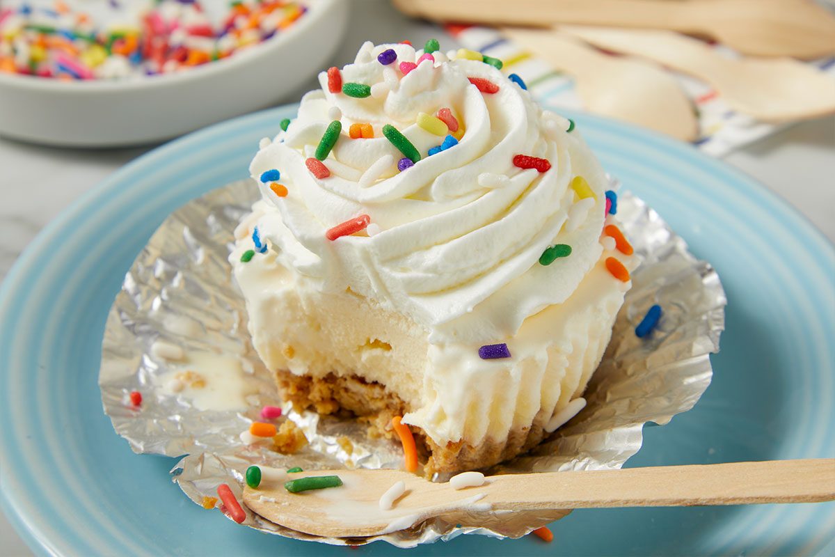 Eye Level horizontal image of a partially eaten mini cheesecake topped with a swirl of white whipped cream and colorful rainbow sprinkles, served on a light blue plate. A wooden spoon rests beside the dessert, which is still in its aluminum foil liner, with some sprinkles scattered on the plate. In the blurred background, a white bowl containing more rainbow sprinkles is visible.