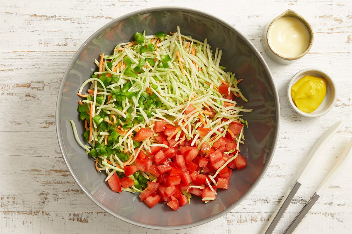 Overhead horizontal image of a gray mixing bowl filled with broccoli coleslaw mix, chopped tomatoes, and chopped jalapeno pepper. Beside the bowl are small dishes holding reduced-fat coleslaw dressing and prepared mustard, with a pair of tongs resting on the wooden surface.