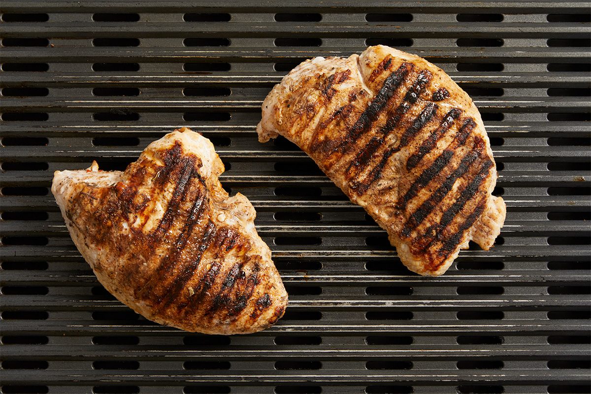 Overhead horizontal image of two turkey breast tenderloins cooking on a griddle, showing prominent dark grill marks on the golden-brown meat. The tenderloins are placed diagonally across the griddle's parallel ridges.