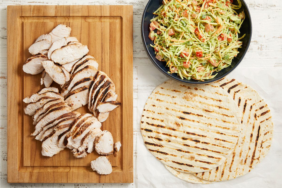 Overhead horizontal image of the components for Jamaican Jerk Turkey Wraps. Sliced grilled turkey breast tenderloins are neatly arranged on a wooden cutting board, positioned next to a bowl of dressed broccoli coleslaw mix containing visible chopped tomatoes and carrots. Two grilled flour tortillas are placed on white parchment paper, ready to be assembled.