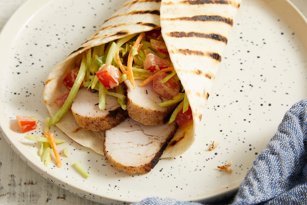 Eye Level horizontal image of a Jamaican Jerk Turkey Wrap, partially open on a white speckled plate. The grilled flour tortilla holds sliced jerk turkey, a vibrant coleslaw mix with broccoli and carrots, and chopped tomatoes.